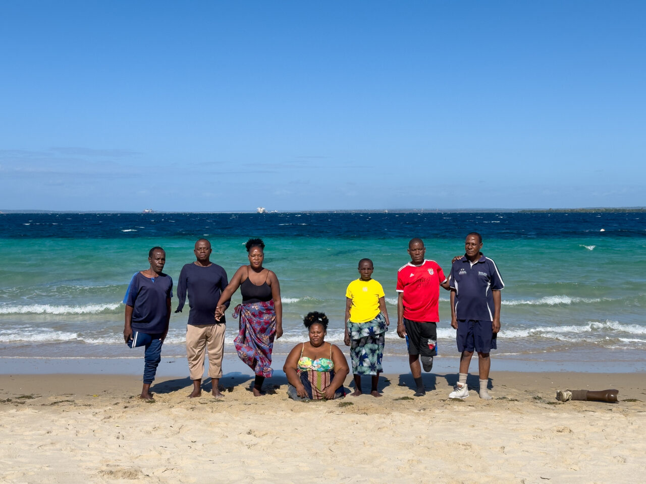Sept personnes amputées sont sur une plage devant la mer. À droite de la photo, une jambe en plastique avec une chaussure de sport est posée sur le sable.