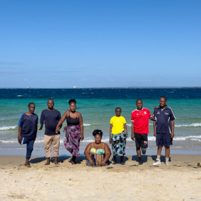 Sept personnes amputées sont sur une plage devant la mer. À droite de la photo, une jambe en plastique avec une chaussure de sport est posée sur le sable.